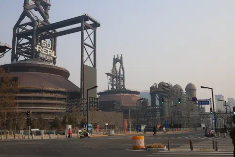 on the side of an old rusty factory building a yellow staircase winds its way up to the top. On the road, a lone person walks past the building.