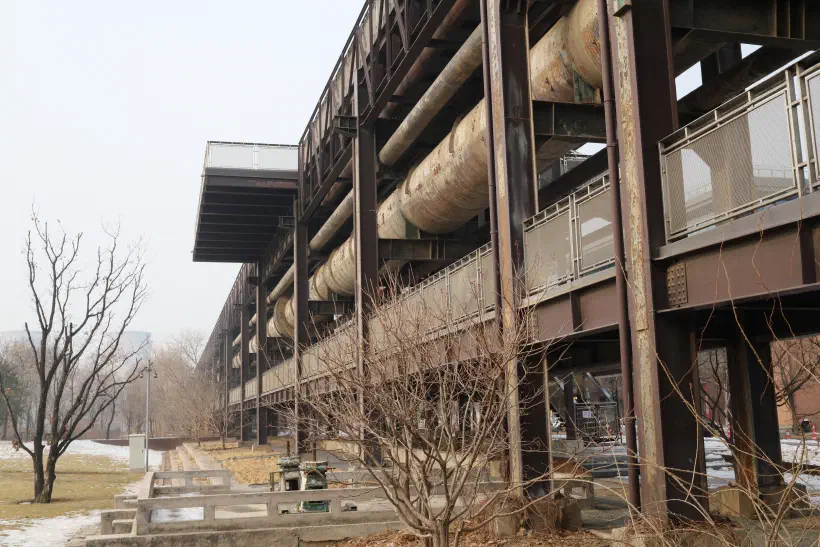 A photo of large metal pipework carried by steel structures into the distance. A walkway hangs underneath the pipes.