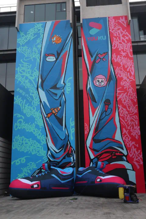 Two strips of canvas hang down the side of a building. One leg is painted on each in a cartoon style. The left leg is predominantly blue, the right leg predominantly red. At the base of each leg, two giant shoes in similar colours.