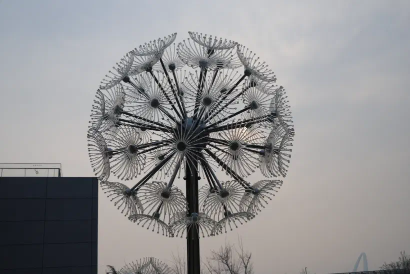 A metal sculpture shaped like a dandilion ready to shed its seeds stands next to a concrete building.