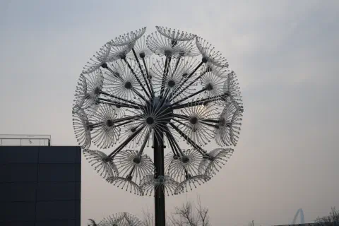 A metal sculpture shaped like a dandilion ready to shed its seeds stands next to a concrete building.