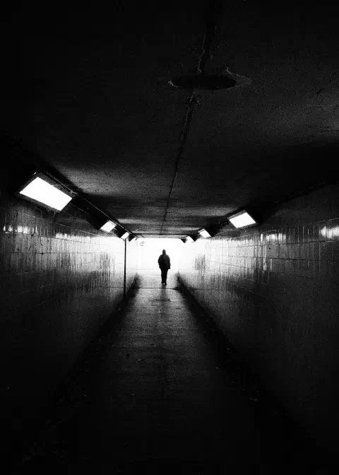A black and white photo taken in an underpass. The light at the far end silhouettes a figure walking down the centre of the tunnel.