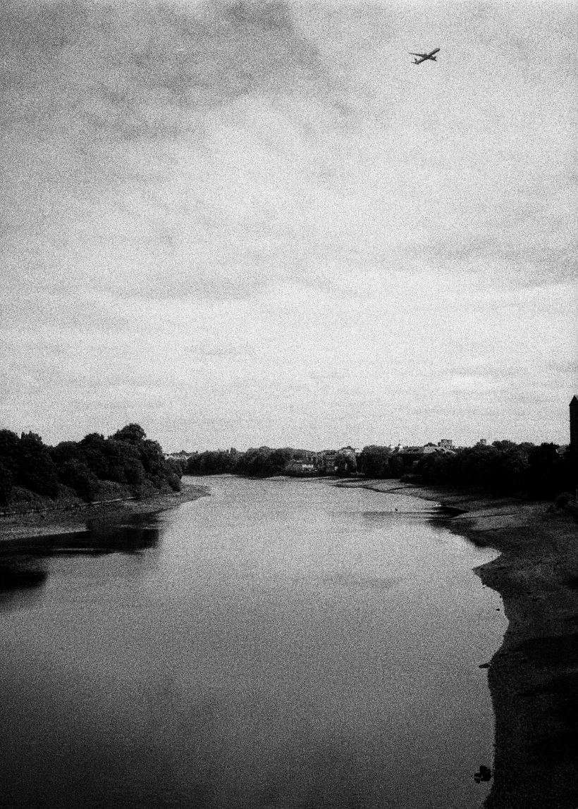 A grainy black and white photo looking along a section of the River Thames in London. The image is taken from a bridge looking over to the river. The water is low enough that you can see the riverbed on the near bank. As you look at the image, the river curves around to the left. In the sky above the river a plane climbs to the right.