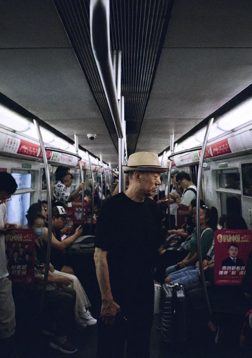 A photograph taken inside a subway train in Beijing. In the centre of the carriage stands a man looking off to the right of the image. We can't see what he is looking at. He appears to be smiling.