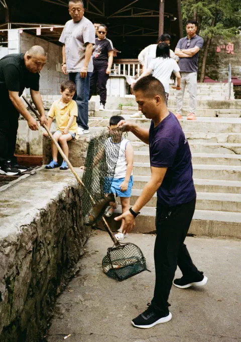A crowd of people including two young boys look on as a man lowers a net containing a freshly caught fish to the floor. Another man reaches out to help take the net off him.