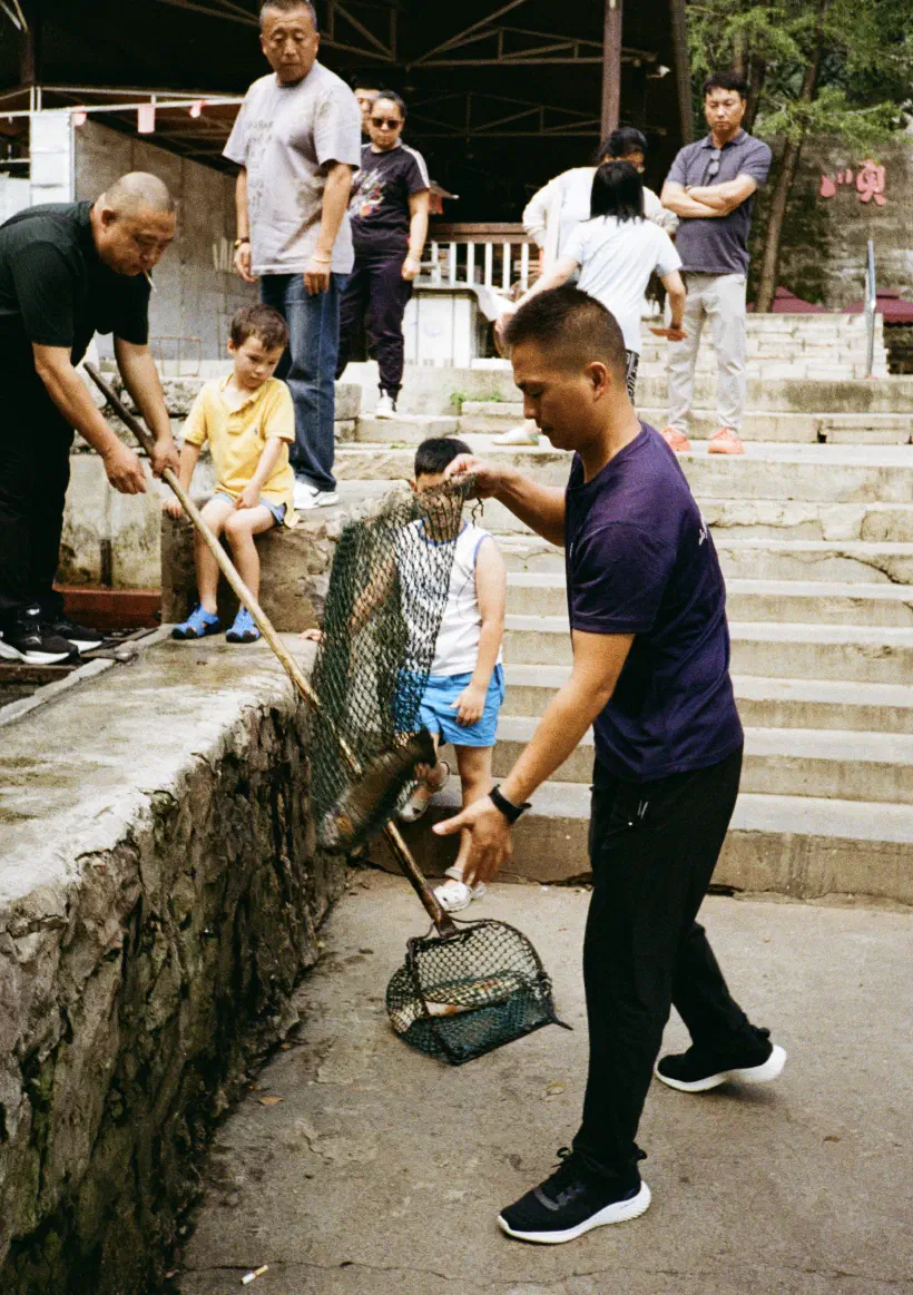 A crowd of people including two young boys look on as a man lowers a net containing a freshly caught fish to the floor. Another man reaches out to help take the net off him.