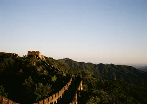 A colour photo of the Great Wall of China. The photograph looks alone a walkable section of the wall which curves round and upwards to a turret on the left of the frame. To the right, green tree covered mountains fall away into the distance.