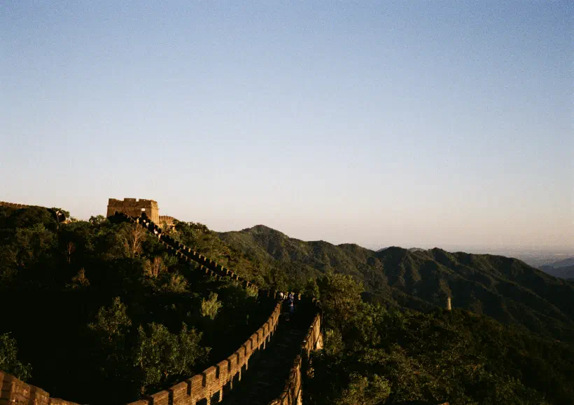 A colour photo of the Great Wall of China. The photograph looks alone a walkable section of the wall which curves round and upwards to a turret on the left of the frame. To the right, green tree covered mountains fall away into the distance.