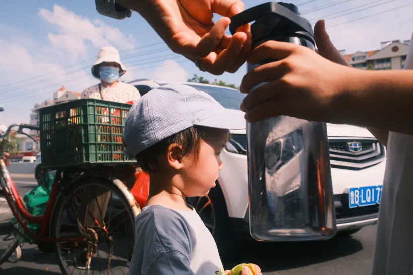 A boy looking out into the road. In his hand he is holding something. Behind him a car and a bicycle, in the foreground a water bottle