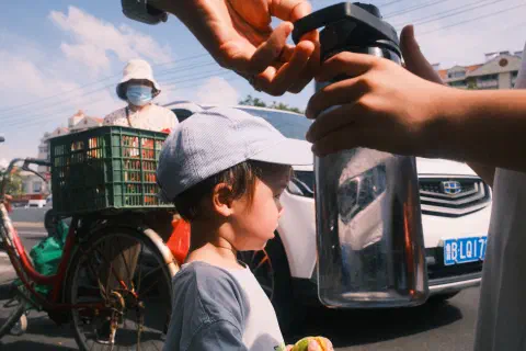 A boy looking out into the road. In his hand he is holding something. Behind him a car and a bicycle, in the foreground a water bottle