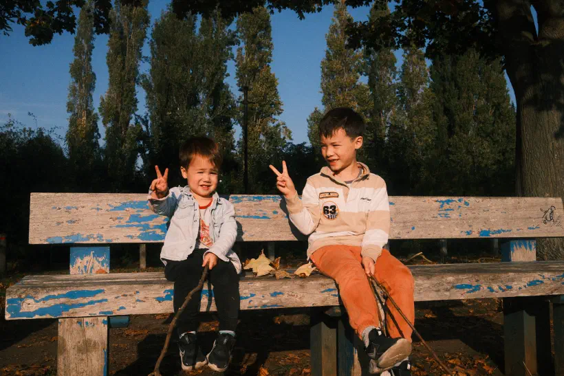 Two boys sit on a public bench making the peace symbol with their hands. With the sun in their eyes, both boys are squinting. Behind them tall trees break up the blue sky.