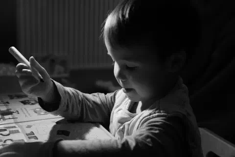 A black and white photo of a young boy sat at a desk holding a pen. He is looking down at a book in front of him. The pen is held high, ready to write.