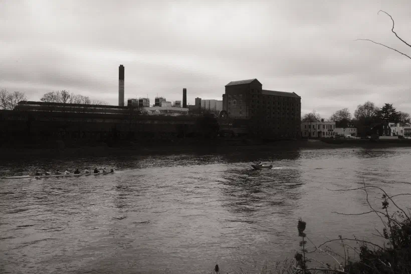 A black and white photo looking out over a river. A rowing boat and safety boat occupy the middle of the river. On the far bank an industrial building and chimney stack mark the skyline.