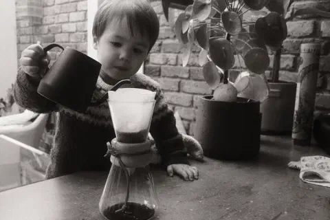 a black and white photo of a boy pouring water into a filter coffee