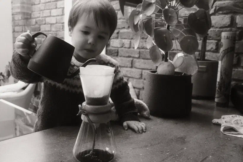 a black and white photo of a boy pouring water into a filter coffee