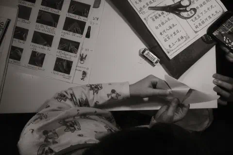 A black and white photo looking down on a child's desk. The child is folding origami. On the desk are a series of instructions in Chinese.