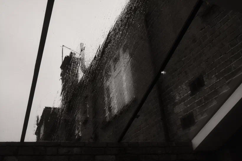 A black and white photo looking up through a glass roof at a brick wall. It's raining outside.