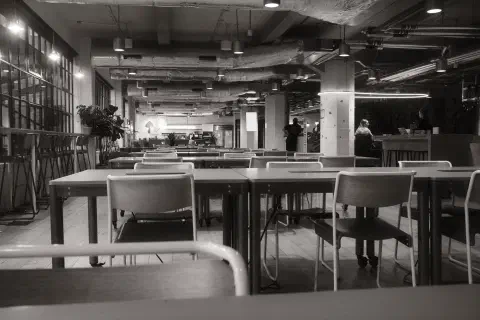 A black and white photo of a empty open plan office. Rows of tables are aligned horizontally across the image with one chair at each desk. The desks are completely empty.