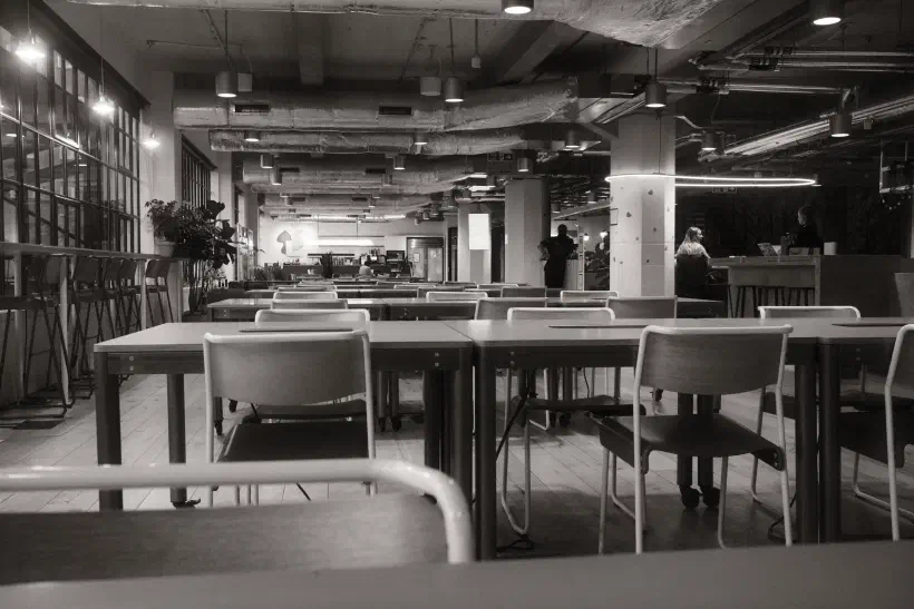 A black and white photo of a empty open plan office. Rows of tables are aligned horizontally across the image with one chair at each desk. The desks are completely empty.