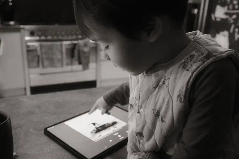 A black and white photo of a young child pointing at an iPad. On the iPad is the cover of 1975's Being Funny in a Foreign Language.