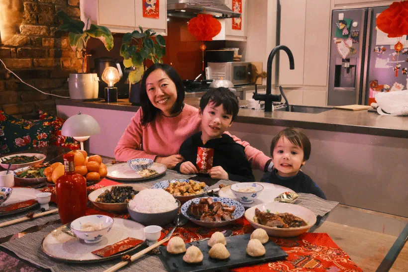A mother and her two boys sit at a table covered in food. One boy is holding a red packet. Red lanterns hang in the background.