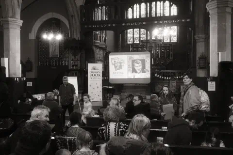 A black and white photo of Lo Cole, author, speaking at Chiswick Book Festival. The festival takes place inside a church.