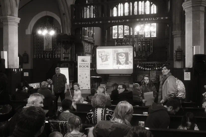 A black and white photo of Lo Cole, author, speaking at Chiswick Book Festival. The festival takes place inside a church.