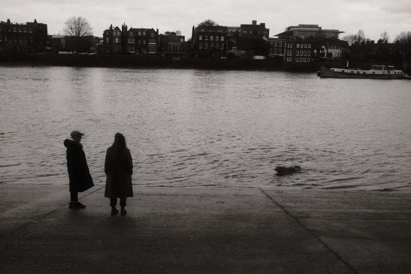 Two people stand on a slipway silhouetted against the river. A dog swims towards them. At the top of the image, houses line the opposite bank of the river.