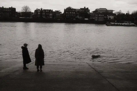 Two people stand on a slipway silhouetted against the river. A dog swims towards them. At the top of the image, houses line the opposite bank of the river.