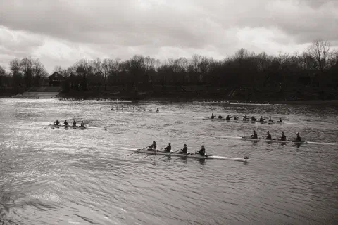 A black and white photo of a river. 9 crews jostle for position before the start of the race. The quads are closer to the bottom of the frame, the eights line up on the far side of the river.