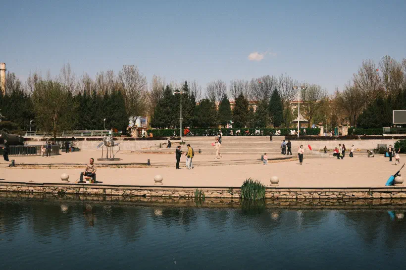 A photo looking over a small river to a paved area on the opposite bank. Families can be seen enjoying the sun; talking, fishing, flying kites, holding balloons.