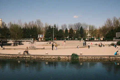 A photo looking over a small river to a paved area on the opposite bank. Families can be seen enjoying the sun; talking, fishing, flying kites, holding balloons.