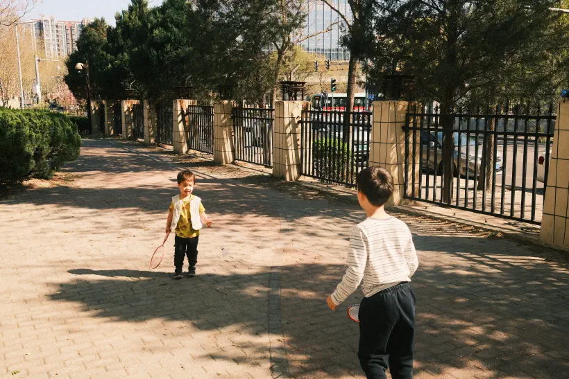 Two boys play badminton together on a paved area fenced off from a road.