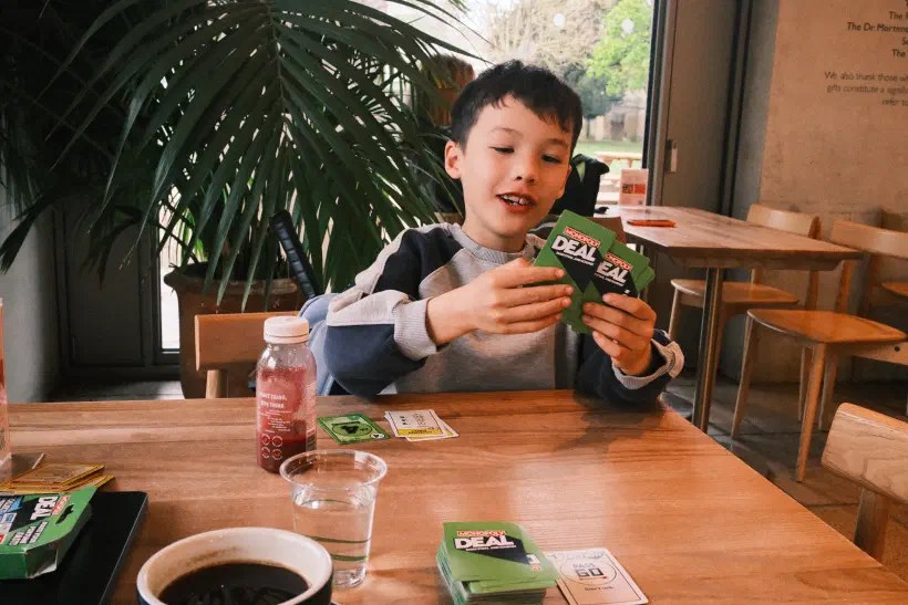 A boy sits at a cafe table holding a number of monopoly deal cards in his hand