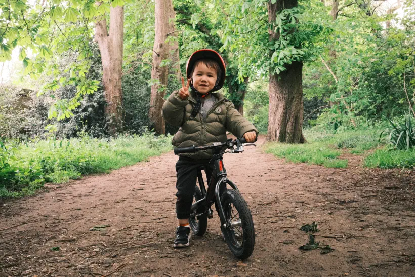 a young boy sits proudly on his balance bike on a dirt trail, trees in the background