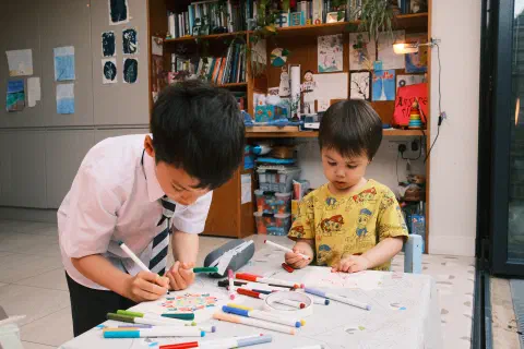 two boys hunched over a small table making greetings cards, the table covered in felt pens