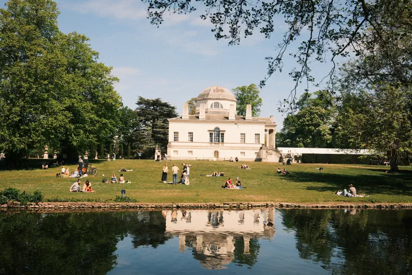 a view over the lake towards Chiswick House, people sat on the lawn enjoying the sunshine