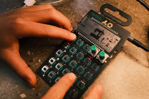 A photo of a Pocket Operator KO by Teenage Engineering. The device looks a bit like a pocket calculator with an array of buttons and an LCD screen. The labels on the buttons are green and two rotary knobs that stick out at the top right of the keys pad are green in colour. Two hands of a young child can be seen pressing the buttons. The child is not visible.