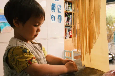 a boy gathers up pieces of dough that have fallen from a pasta machine, strands of pasta hang closes to the camera, a wide sheet of uncut pasta hangs behind them, out of shot the feed through a pasta cutter