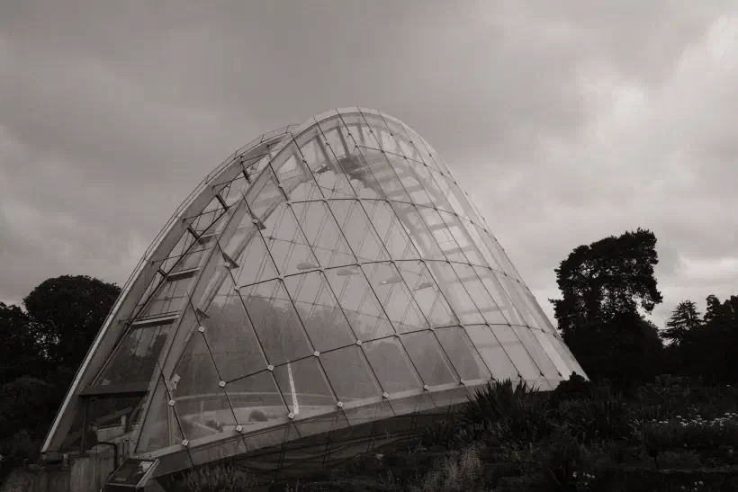 A black and white photo of Alpine House at Kew Gardens, the narrow semicircular building is made of glass and steel.