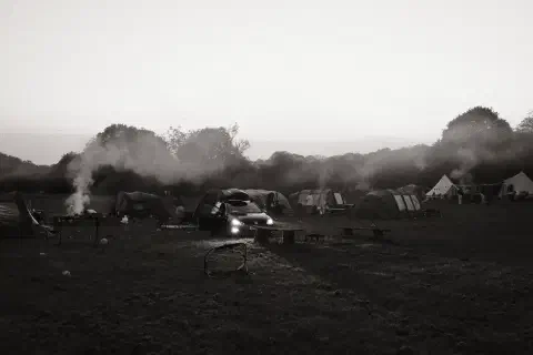 a black and white photo of a campsite, smoke rising from a couple of camp fires, a car with its headlights on faces towards the camera