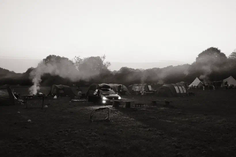 a black and white photo of a campsite, smoke rising from a couple of camp fires, a car with its headlights on faces towards the camera