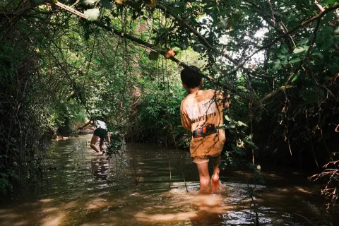a boy follows a girl down a muddy stream, the water up to their ankles, either side of the stream branches and leaves provide shade from the sun
