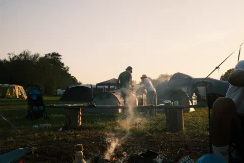 Smoke rises from a camp fire. In the distance, two people stand facing away from the camera looking down at something behind a tent. A few embers of the fire can be seen in the foreground.