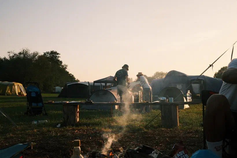Smoke rises from a camp fire. In the distance, two people stand facing away from the camera looking down at something behind a tent. A few embers of the fire can be seen in the foreground.