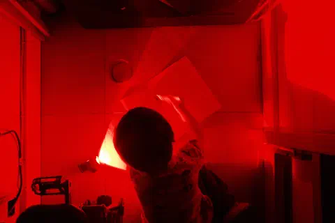 a boy sits on the floor painting light sensitive solution onto a piece of paper, the image is lit by light from a red bike light