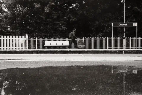A black and white photo of a man walking along the station platform at Chiswick station. In the foreground a pool of water provides a reflection.