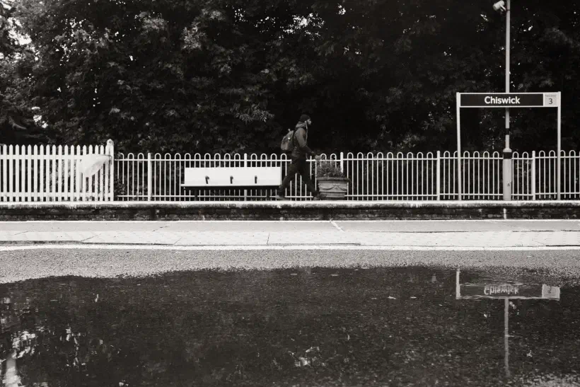 A black and white photo of a man walking along the station platform at Chiswick station. In the foreground a pool of water provides a reflection.