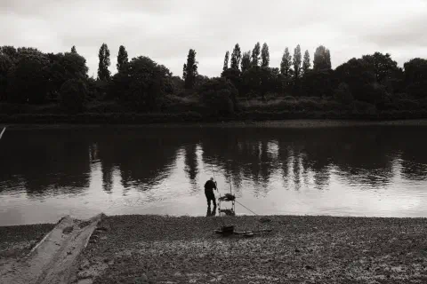 A black and white photo of a man of a man standing at the edge of a river fishing.