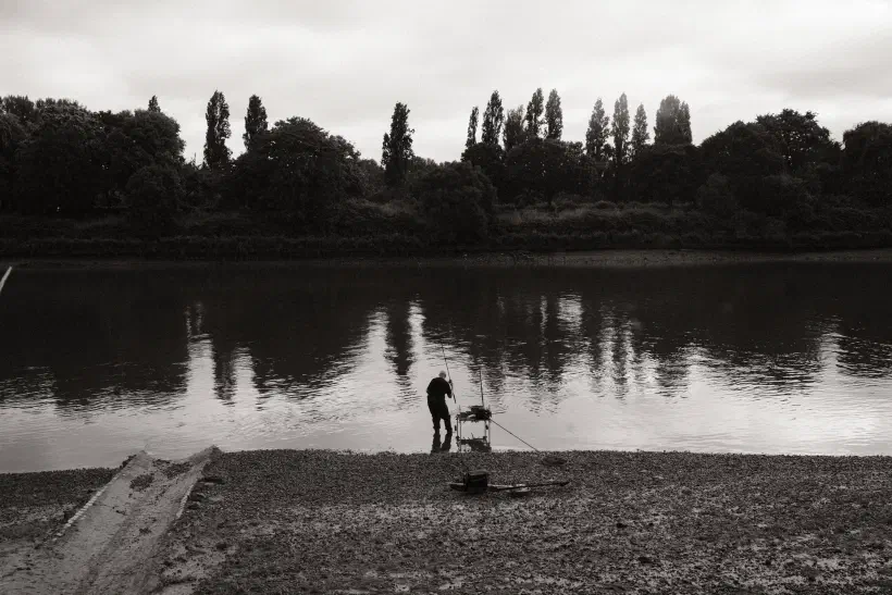 A black and white photo of a man of a man standing at the edge of a river fishing.